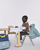 A young child sits at a small wooden desk in a bright studio setting, wearing a brown sweater over a white collared shirt, dark shorts, green socks, and white shoes. A light blue puffer jacket is draped over the chair behind them, and a blue-and-light-blue backpack rests on the floor to the left.