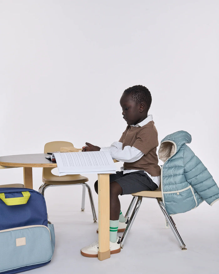 A young child sits at a small wooden desk in a bright studio setting, wearing a brown sweater over a white collared shirt, dark shorts, green socks, and white shoes. A light blue puffer jacket is draped over the chair behind them, and a blue-and-light-blue backpack rests on the floor to the left.