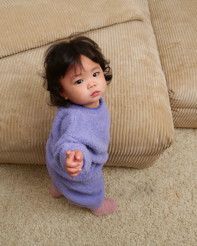 A toddler standing by a beige couch, wearing a soft, fuzzy knit taro purple sweater, pants, and a matching beanie.