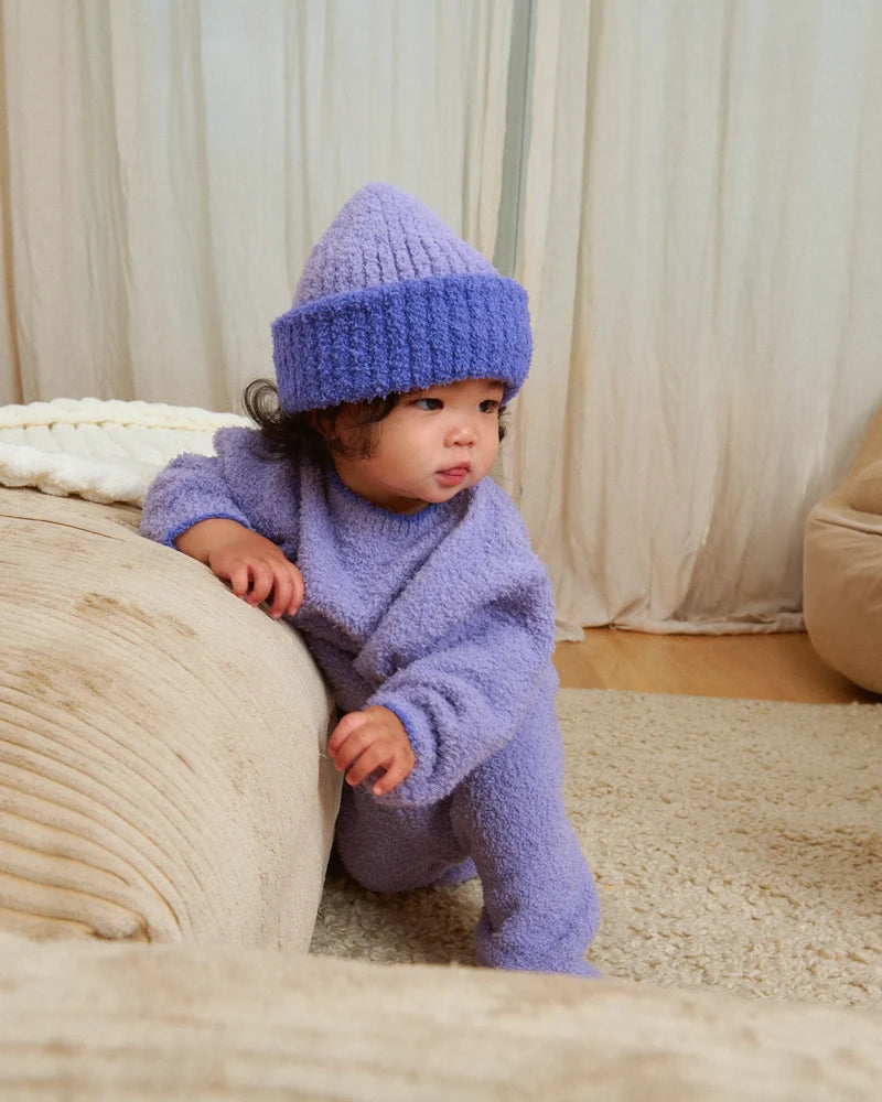 A baby standing by a beige couch, wearing a soft, fuzzy knit taro purple sweater, pants, and a matching beanie.