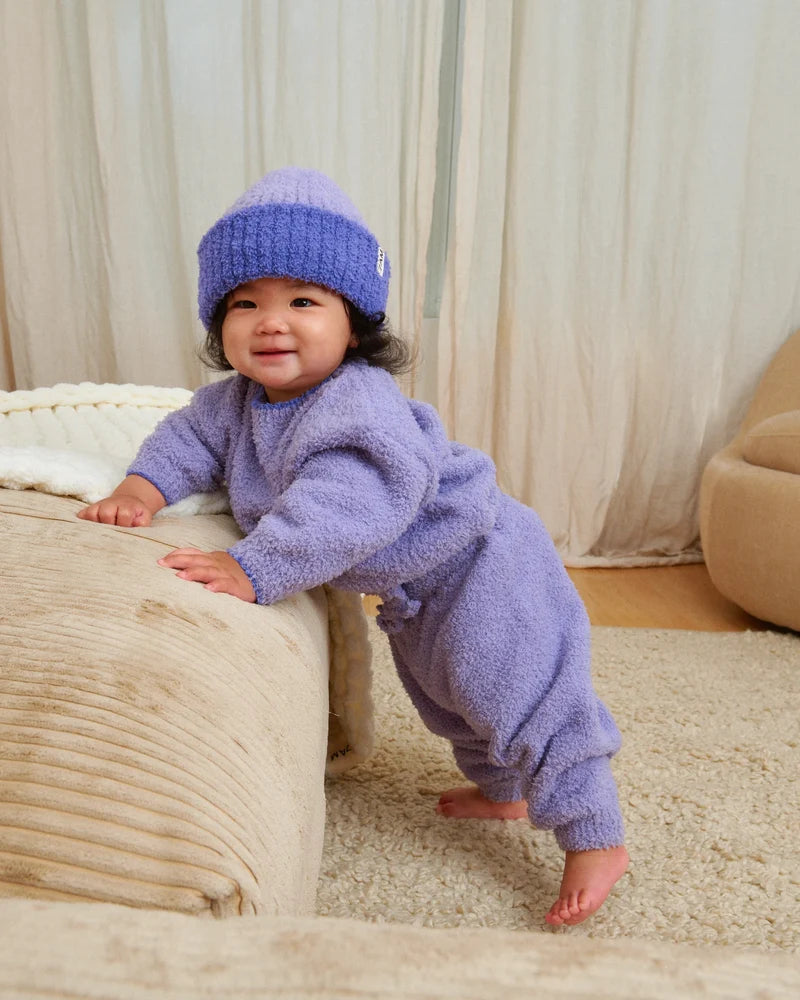 A baby standing by a beige couch, wearing a soft, fuzzy knit taro purple sweater, pants, and a matching beanie.