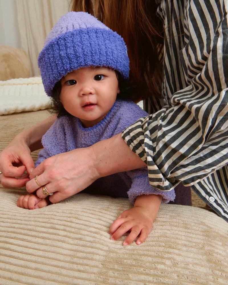 A toddler wearing a matching fuzzy purple knit sweater and a ribbed beanie with a folded brim and a white "7AM" tag.