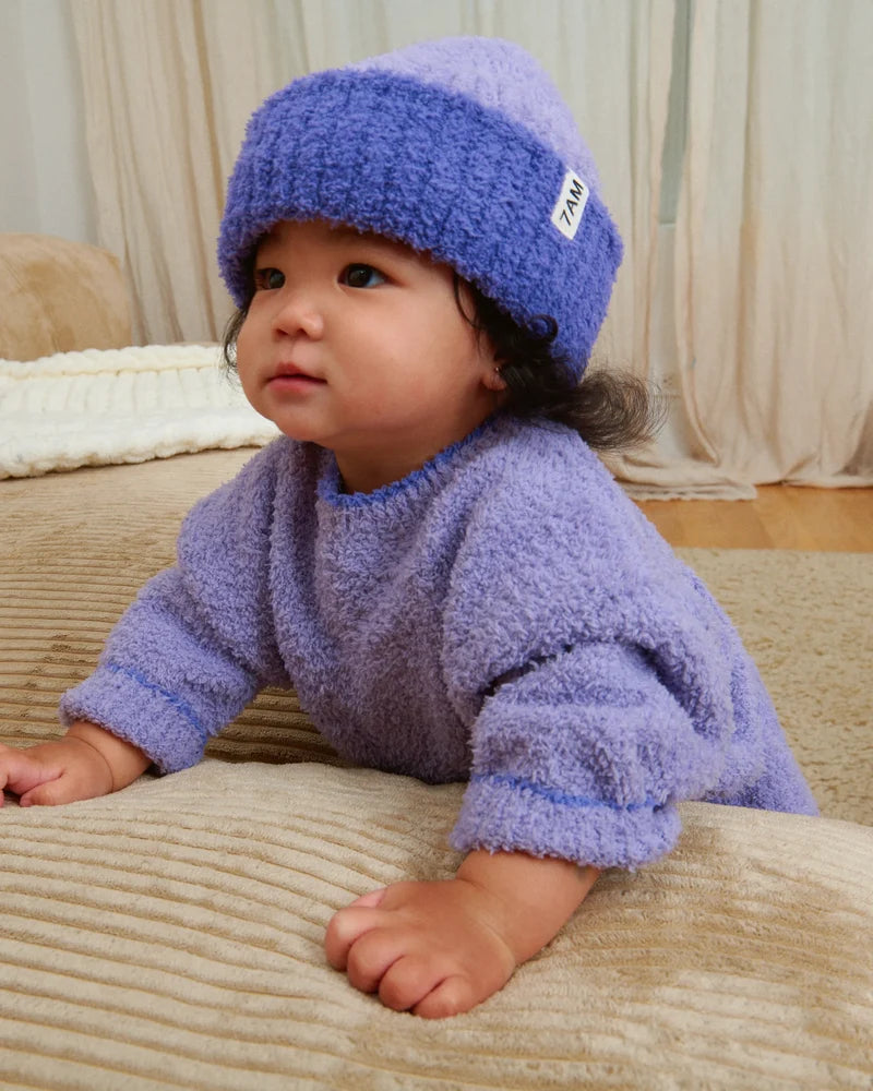A toddler wearing a matching fuzzy purple knit sweater and a ribbed beanie with a folded brim and a white "7AM" tag.