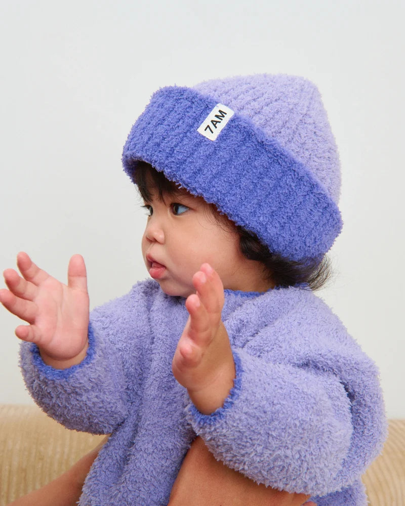 Child wearing a purple fleece outfit with a matching hat, sitting on a beige couch.