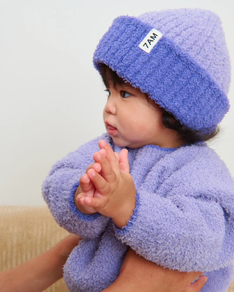 A toddler wearing a matching fuzzy purple knit sweater and a ribbed beanie with a folded brim and a white "7AM" tag.
