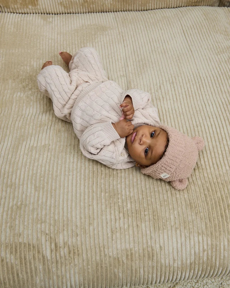 A baby lying on a textured neutral-colored blanket, wearing a matching ribbed cream-colored outfit and a fuzzy pecan-colored knit bear beanie with small ears.