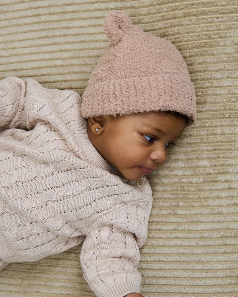 A baby wearing a cream cable-knit sweater and a dusty pink knit beanie hat, lying on a ribbed beige blanket while holding an adult's hand.
