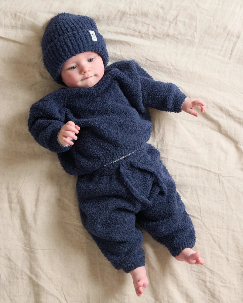 A toddler lying on a light neutral surface, wearing a matching navy blue fuzzy knit long-sleeve top, lounge pants, and a knit beanie.