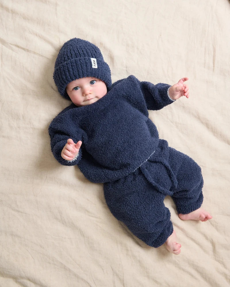 A baby wearing a dark navy blue ribbed knit beanie and a matching fuzzy long-sleeved outfit while lying on a soft, light-colored surface.