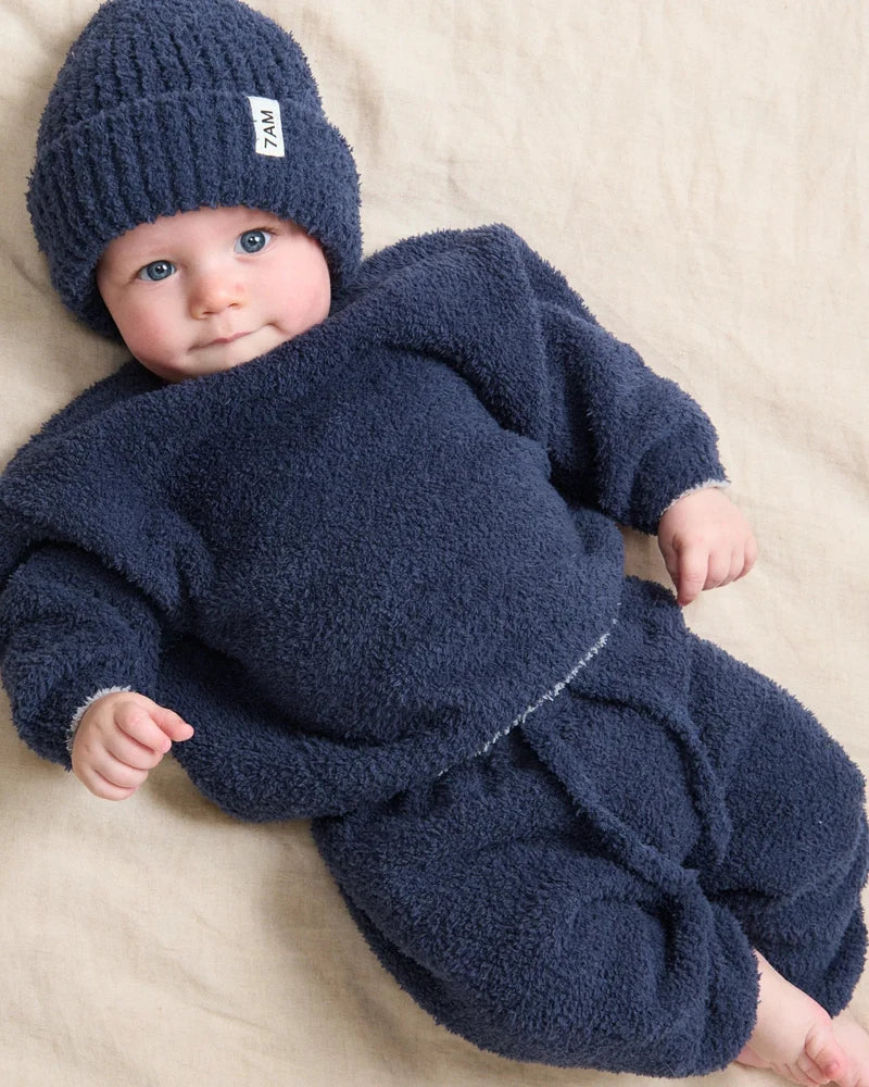 A toddler wearing a matching navy blue fuzzy knit outfit and beanie, lying down against a neutral background.