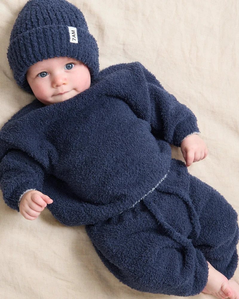 A baby lying down and wearing a dark navy blue ribbed knit beanie with a small white brand tag and a matching fuzzy long-sleeved outfit.