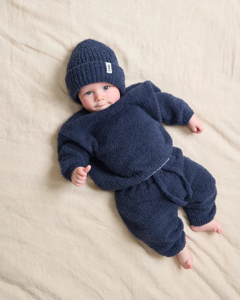 A baby lying on a beige fabric surface wearing a matching navy blue fuzzy knit outfit with a beanie hat. A small white tag on the hat reads 'BEBE'.