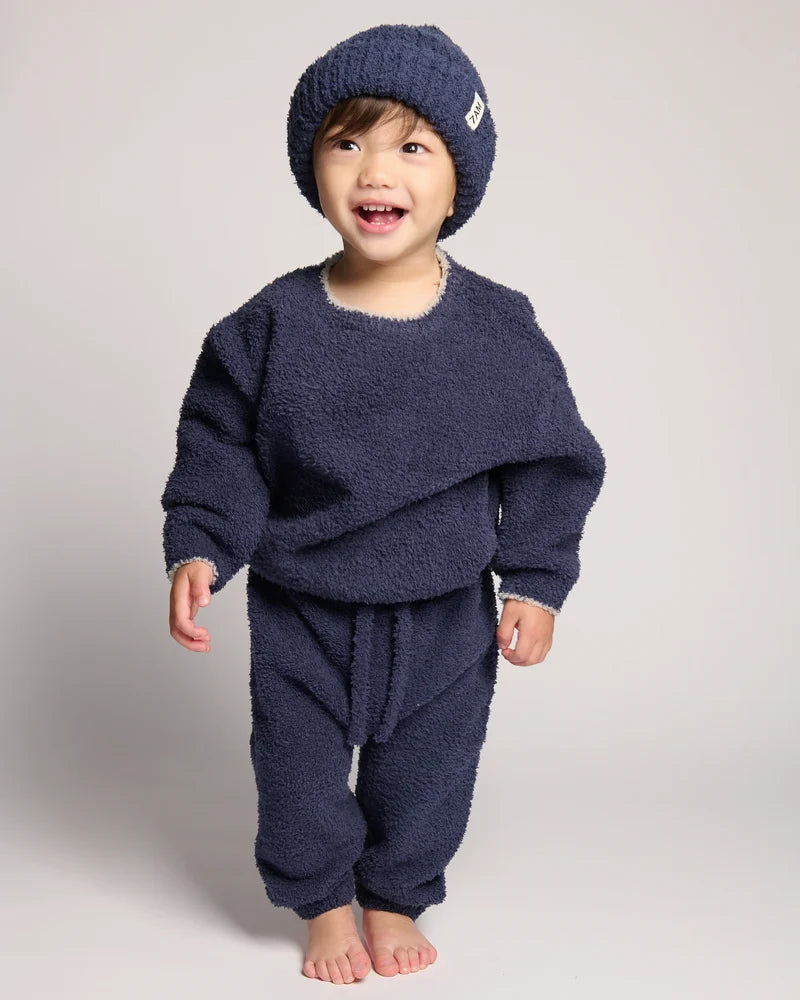 A young child standing while wearing a dark navy blue ribbed knit beanie and a matching fuzzy long-sleeved outfit against a plain background.