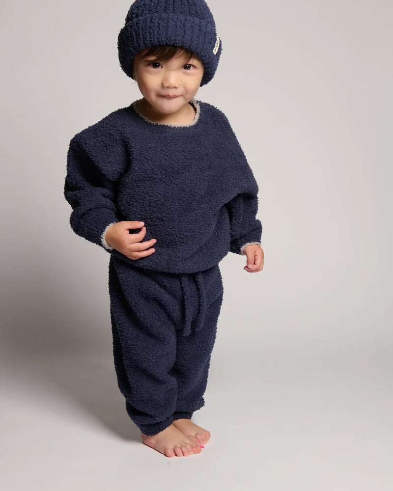 A toddler wearing a cozy navy blue fuzzy knit outfit, including a matching beanie, sweatshirt, and pants, photographed against a neutral light gray studio background.