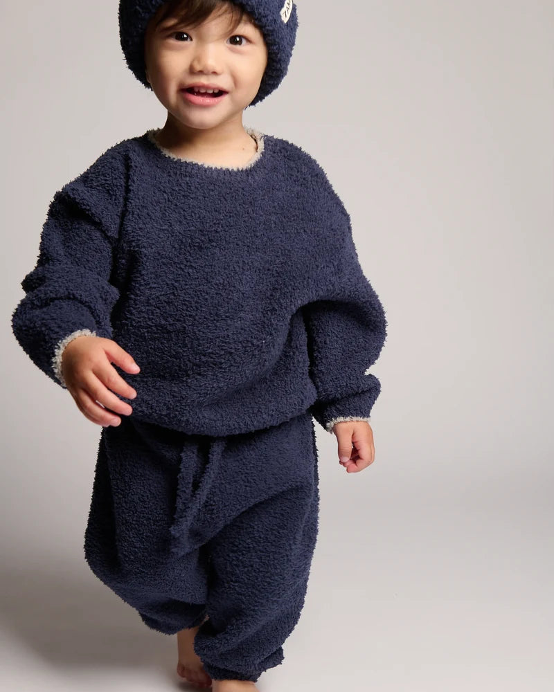 A smiling toddler wearing a matching navy blue fuzzy fleece outfit with a knit beanie, photographed against a plain light gray studio background.