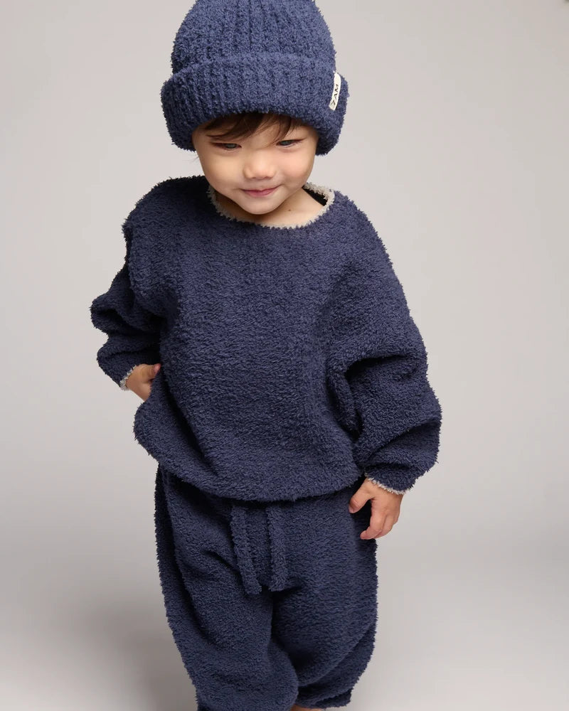 A toddler wearing a cozy navy blue fuzzy knit outfit, including a matching beanie, sweatshirt, and pants, photographed against a neutral light gray studio background.