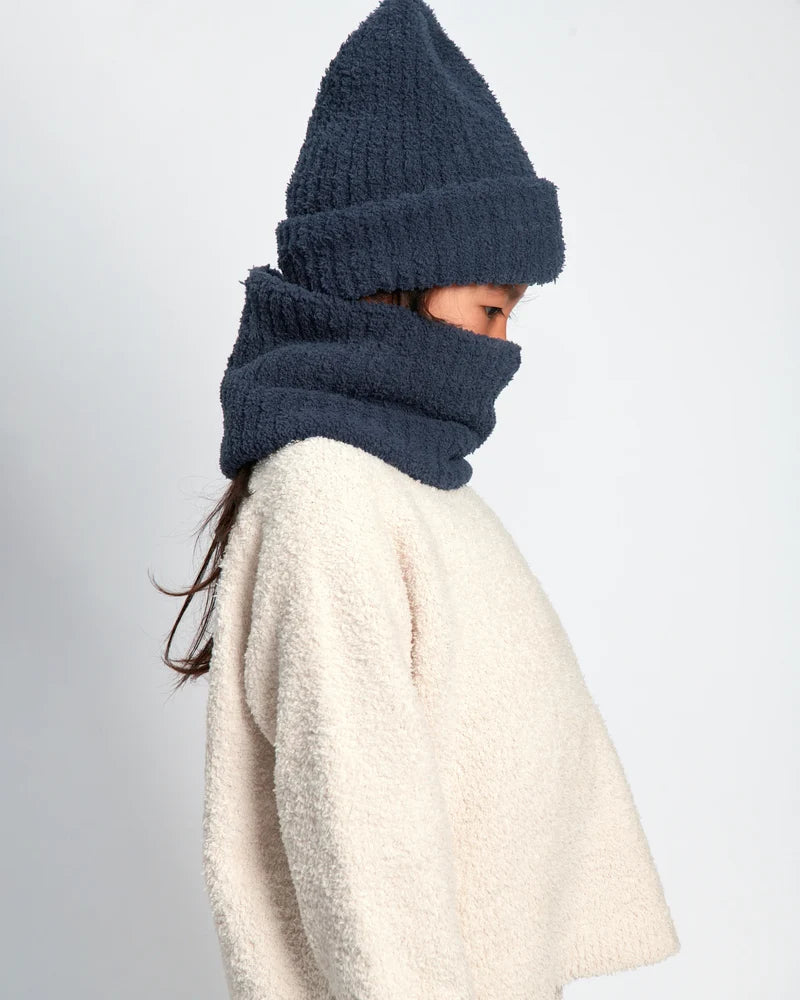 A child wearing a dark navy blue fuzzy knit beanie and matching neck warmer cowl over a cream sherpa sweater, shown in profile against a light gray background.