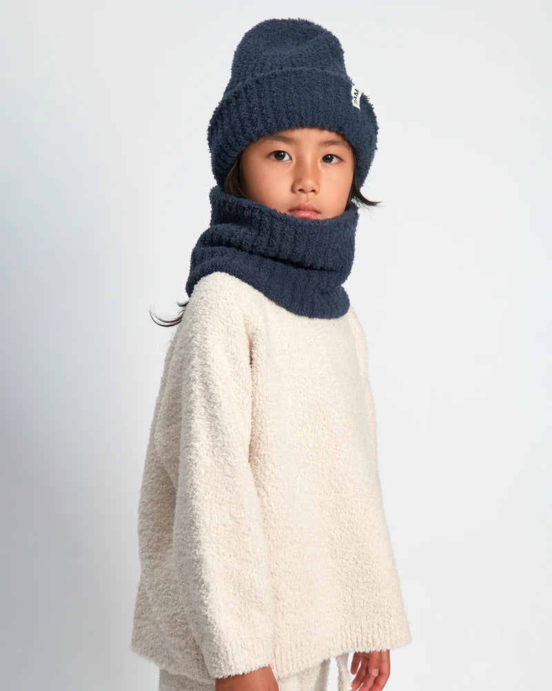 A child wearing a navy blue knit beanie with a small white logo and a matching navy blue knit neck warmer, paired with a cream sherpa sweater, against a light gray studio background.