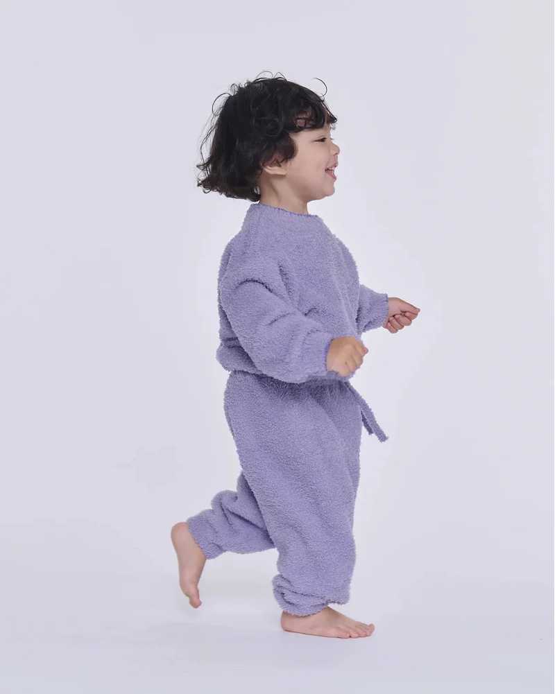 A toddler walking sideways against a light gray background, wearing a fuzzy lavender long-sleeve sweatshirt and matching lounge pants.
