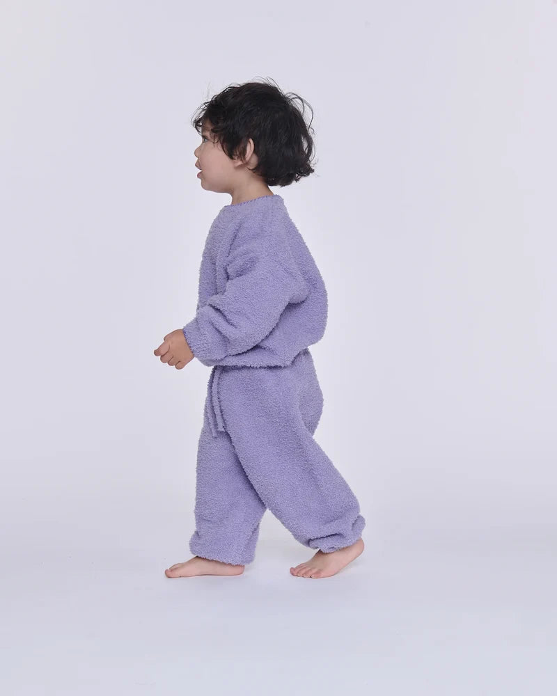 A toddler walking sideways against a light gray background, wearing a fuzzy lavender long-sleeve sweatshirt and matching lounge pants.