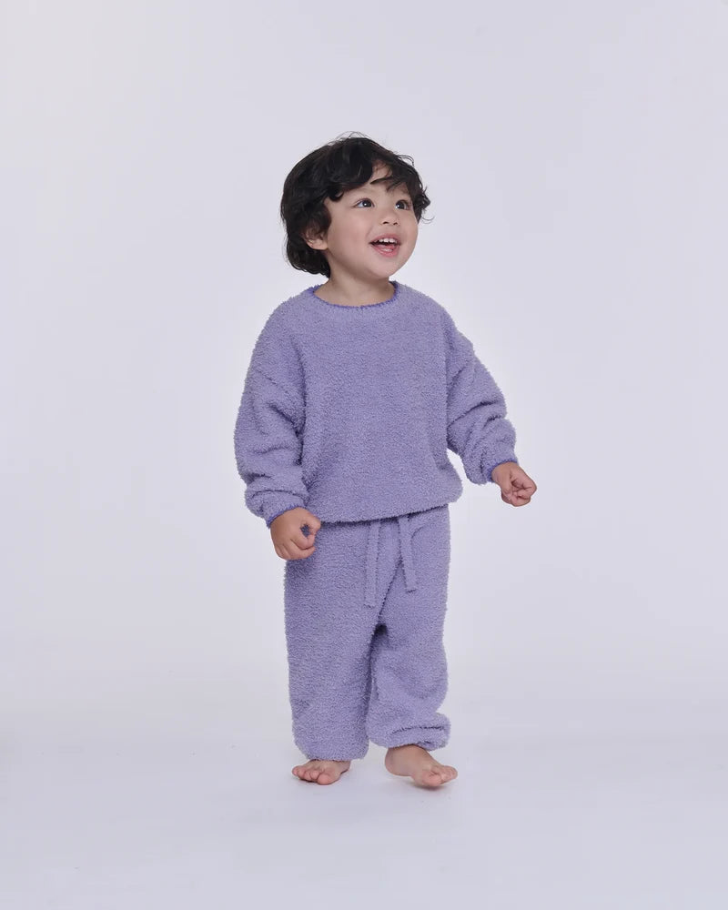 A toddler standing barefoot against a light gray studio background, wearing a fuzzy lavender long-sleeve top and matching drawstring pants.