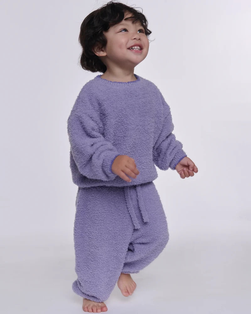 A toddler wearing a fuzzy lavender two-piece loungewear set, consisting of a long-sleeve sweatshirt and matching pants, standing against a plain light gray background.