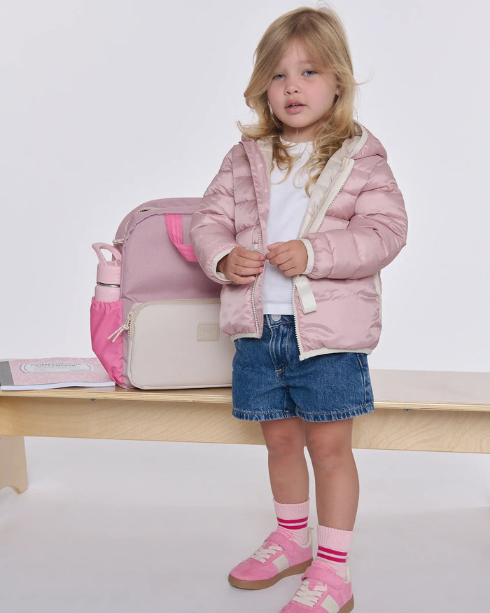 A young girl stands on a wooden bench wearing a light pink puffer jacket, blue denim shorts, pink sneakers, and pink striped socks, with a matching pink and cream backpack beside her.