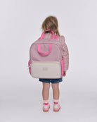 A young child viewed from behind wearing a dusty pink puffer jacket, denim shorts, and pink shoes, carrying a large structured diaper bag backpack in dusty pink and cream. The backpack features bright pink handles, side pockets, and a cream front pocket with a small metallic brand plaque.