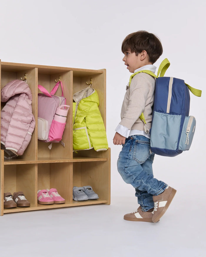A young child wearing a beige long-sleeve shirt, blue jeans, and brown shoes walks past a wooden cubby shelf while wearing a blue and light blue backpack with lime green straps. The cubbies contain various children's items like jackets and shoes against a clean white background.