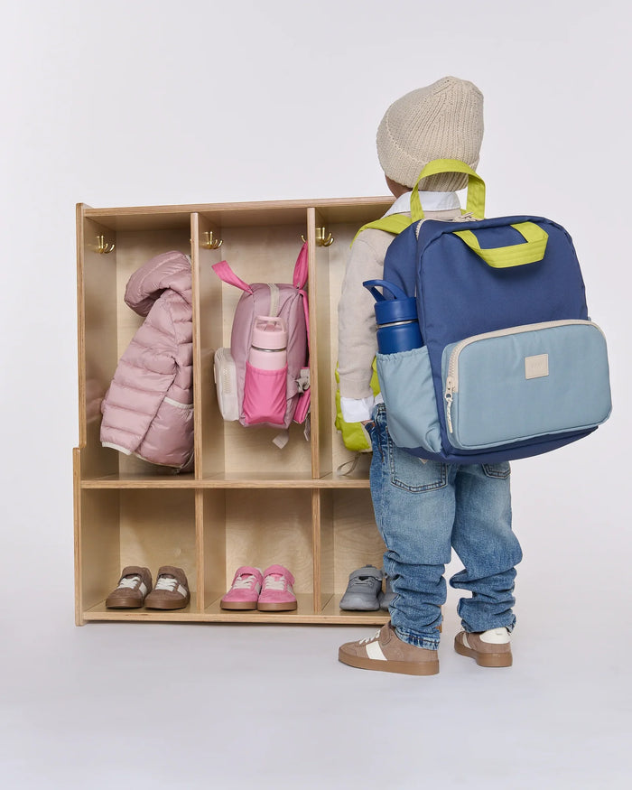 Child with a blue backpack standing next to a wooden locker with children's items inside.
