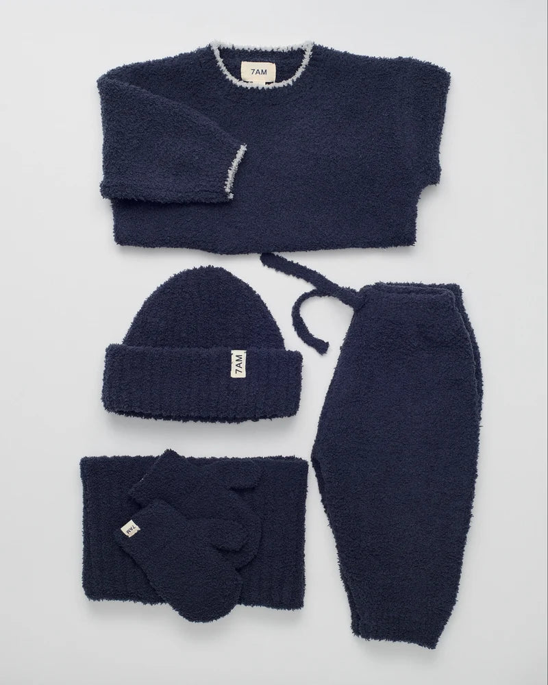 A dark navy blue knit loungewear set laid out on a light gray background, including a crewneck sweater, a beanie, sweatpants, a scarf, and gloves. The knit fabric has a soft, fuzzy texture, and each item features a small white brand tag with 'YAKI' printed on it.