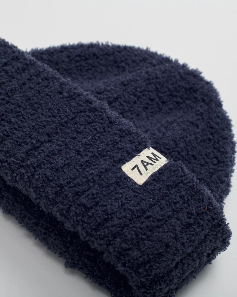 A close-up of a dark navy blue fuzzy fleece beanie with a small white rectangular label reading '7AM' on the cuff, set against a light gray background.