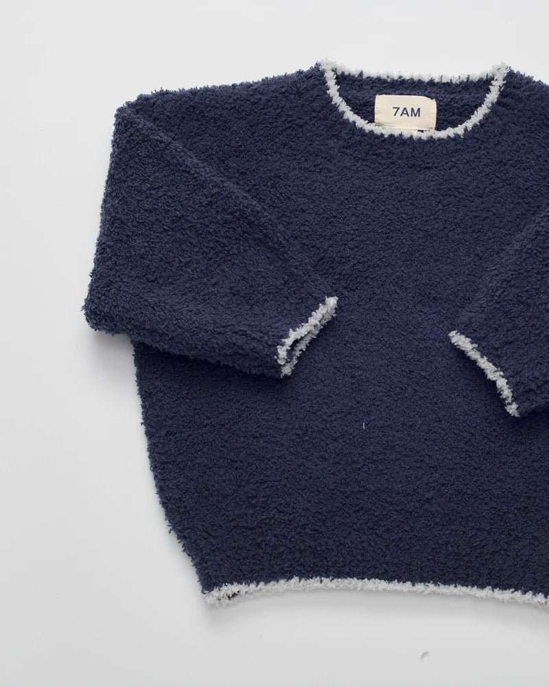 A navy blue fuzzy long-sleeve sweater with light gray trim, laid flat on a white background. The brand label inside the collar reads '7AM'.