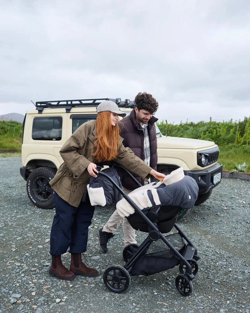 A woman pushes a stroller with a baby inside while walking outdoors next to a man in front of a beige off-road vehicle under an overcast sky, with the baby carriage outfitted with a Benji Applique Car Seat Cocoon by 7AM Enfant.