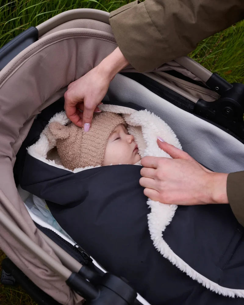 An adult adjusting a stroller bag with white fleece lining while a baby wearing a bear-eared knit beanie rests inside the stroller.