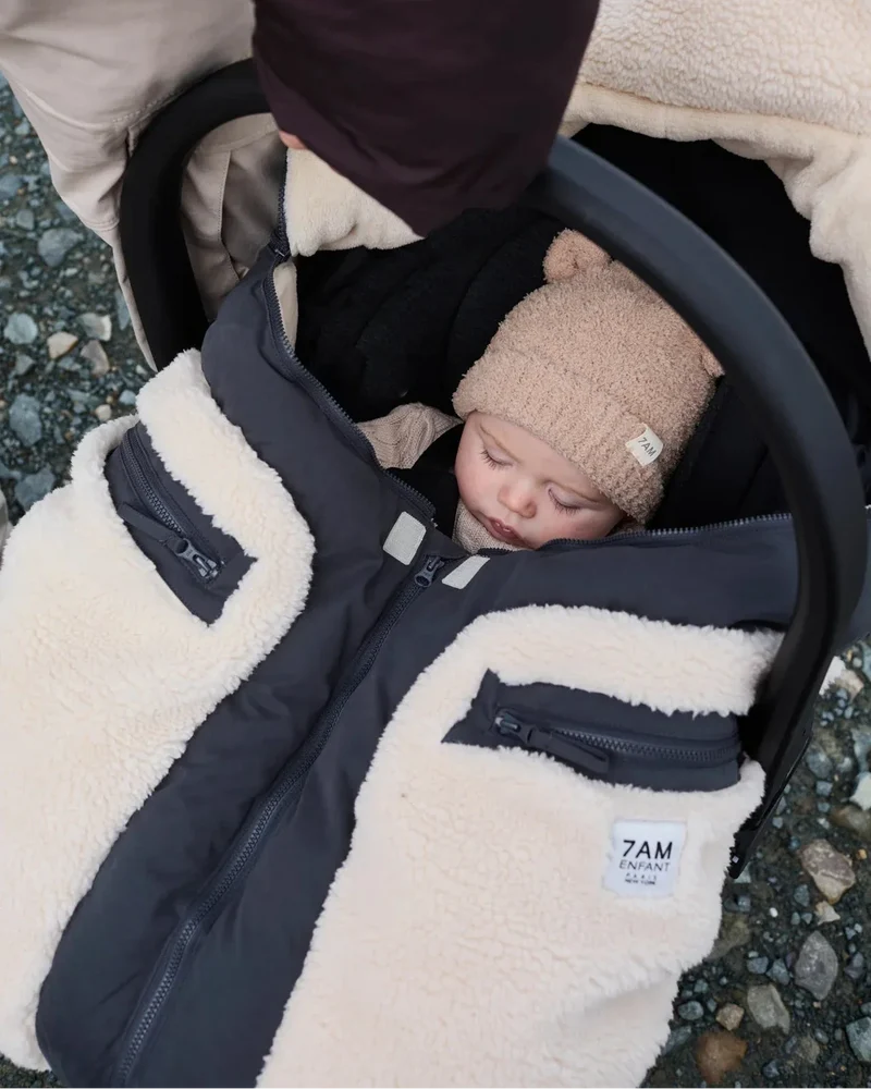 A baby sleeping in a black stroller wearing a beige knit beanie, covered by a cream and black fleece-lined stroller bag with zippered pockets. A small white label on the bag reads '7AM ENGLAND'.