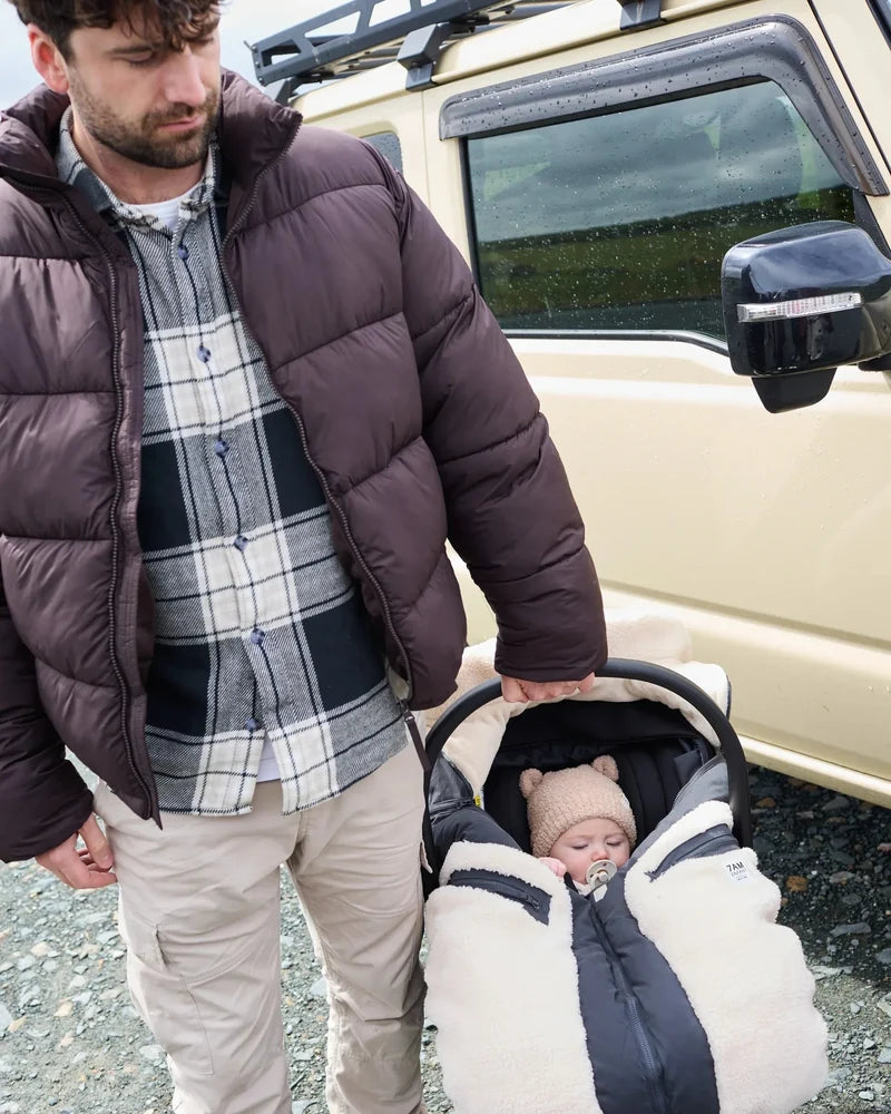 A man standing next to an SUV outdoors, holding a baby in a stroller who is wearing a fuzzy bear-eared beanie and a warm winter bunting bag.