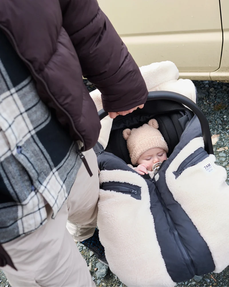 A baby wearing a beanie and pacifier is snuggled inside an infant car seat with the Benji Applique Car Seat Cocoon by 7AM Enfant featuring a dark navy or black outer shell and ivory sherpa trim, while a person stands nearby holding the car seat handle.