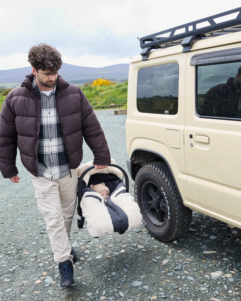 A man walking beside a cream-colored off-road vehicle, carrying a baby in a padded car seat. The man wears a dark brown puffer jacket over a plaid shirt and beige pants, set against a gravel ground and distant hills.