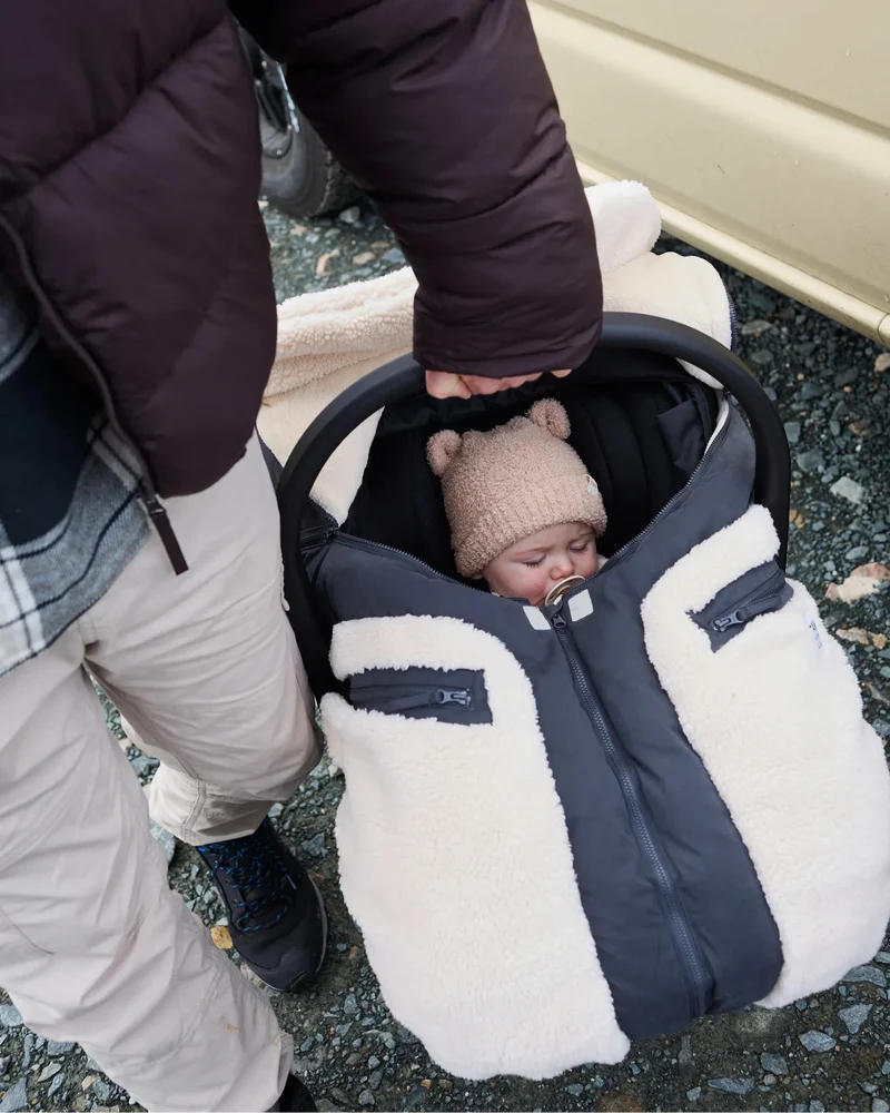 A baby sleeping in a stroller wearing a beige knit beanie, covered by a cream and black fleece-lined stroller bag with zippered pockets.