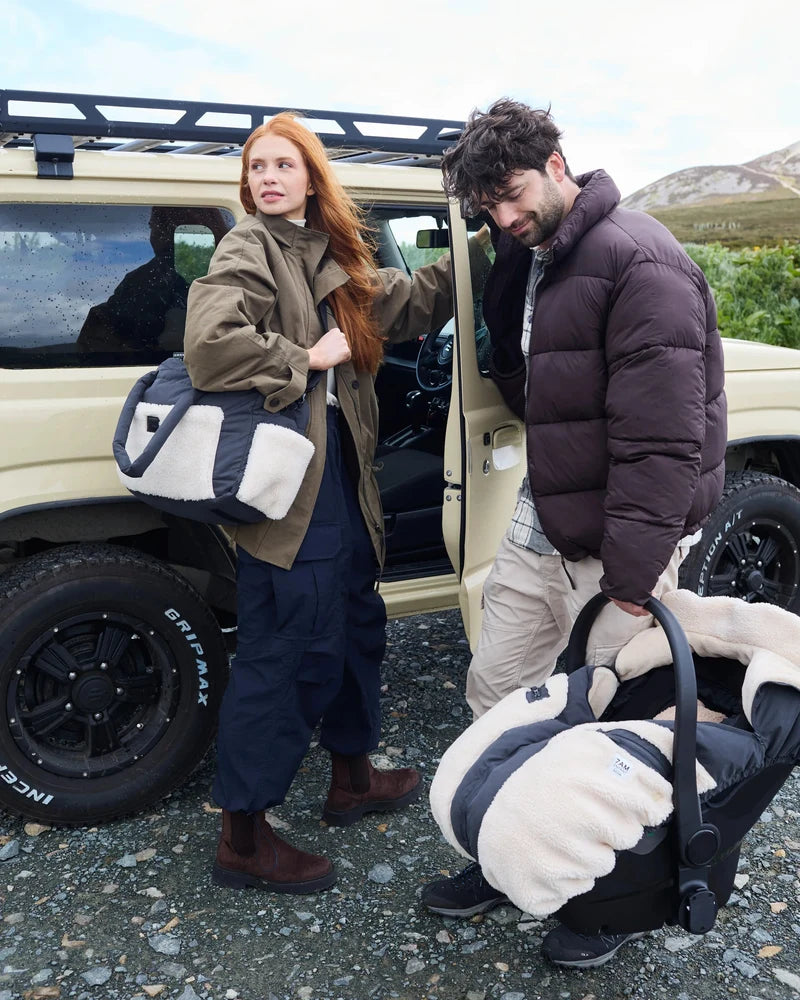 A man and a woman outdoors beside a beige off-road vehicle, each holding a matching black and cream fleece car seat liner set. The woman wears an olive green jacket and navy pants, and the man wears a dark brown puffer jacket and khaki pants.