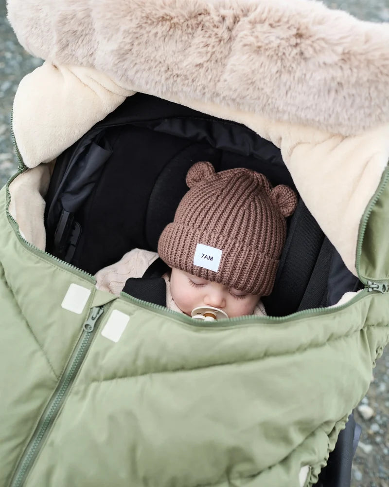 Baby sleeping peacefully in a moss brown Helsinki Car Seat Cocoon by 7AM Enfant with a plush faux-fur collar and matching knit bear-ear beanie.