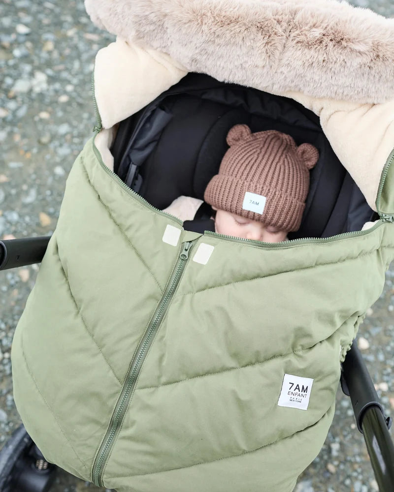 Baby sleeping peacefully in a moss brown Helsinki Car Seat Cocoon by 7AM Enfant with a plush faux-fur collar and matching knit bear-ear beanie.