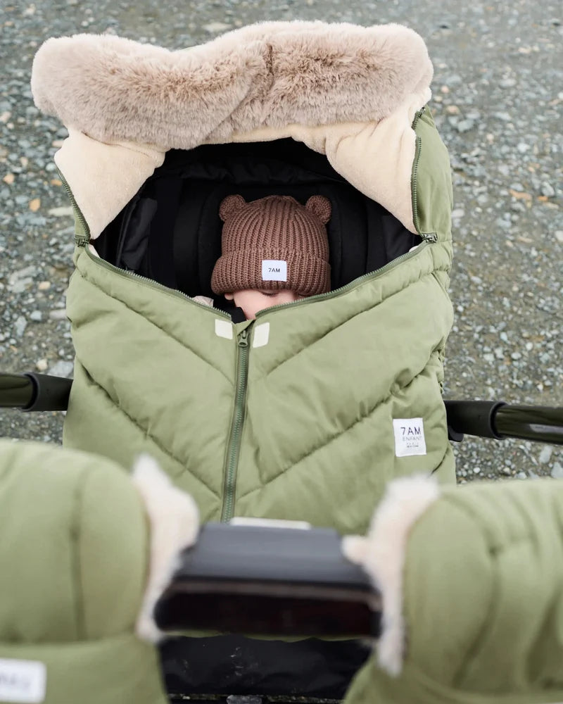 Baby sleeping peacefully in a moss brown Helsinki Car Seat Cocoon by 7AM Enfant with a plush faux-fur collar and matching knit bear-ear beanie.