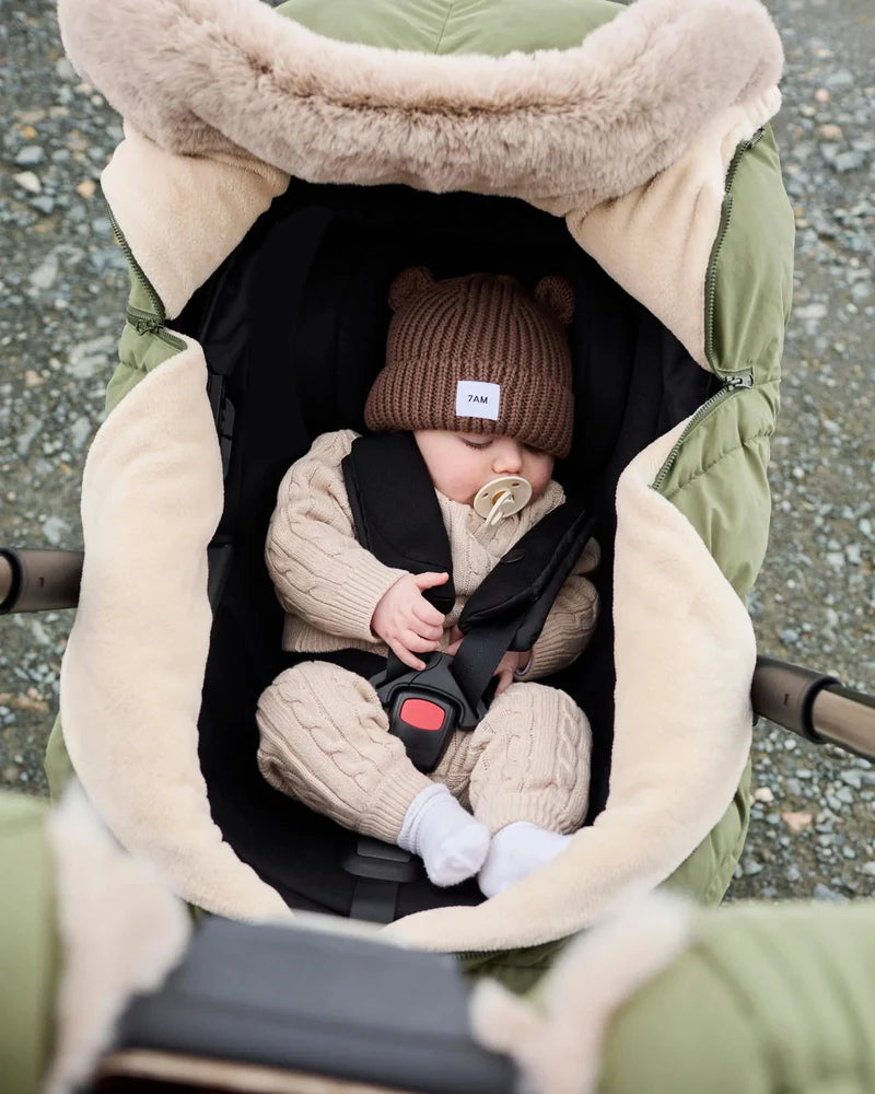 Baby sleeping peacefully in a moss brown Helsinki Car Seat Cocoon by 7AM Enfant with a plush faux-fur collar and matching knit bear-ear beanie.