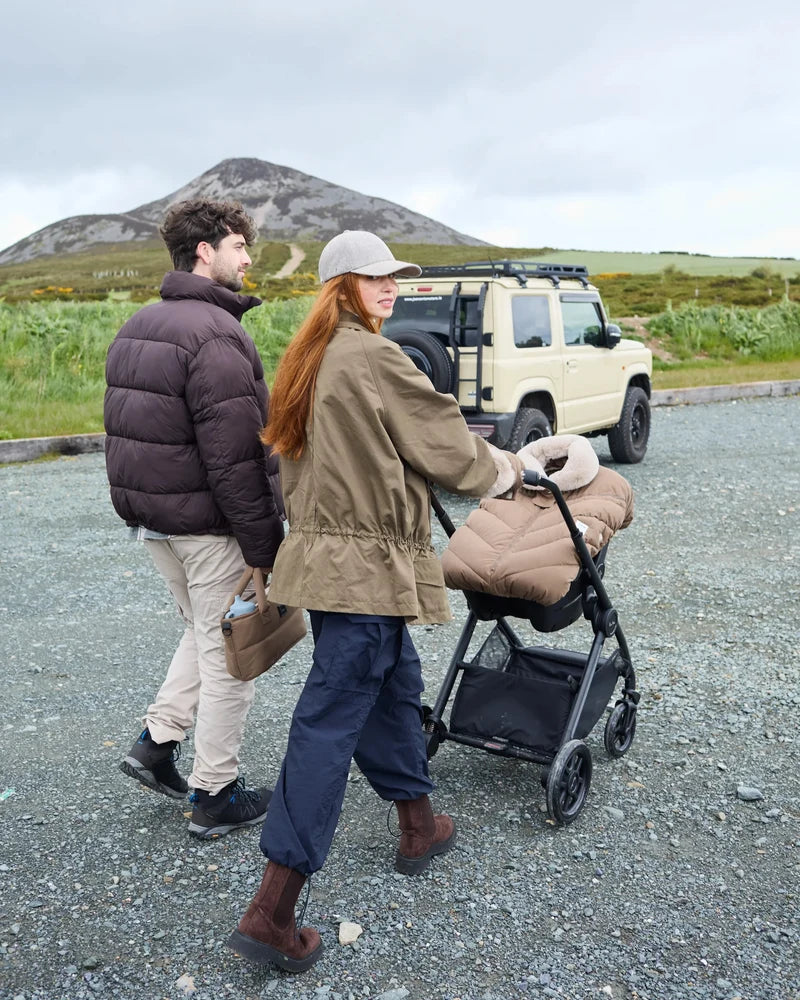Lifestyle view of parents outdoors with a stroller featuring the moss brown Helsinki Car Seat Cocoon by 7AM Enfant parked near an off-road vehicle with a mountain landscape in the background.