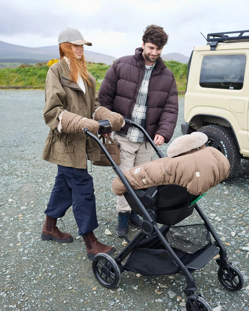 Lifestyle view of parents outdoors with a stroller featuring the moss brown Helsinki Car Seat Cocoon by 7AM Enfant parked near an off-road vehicle with a mountain landscape in the background.