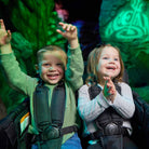 Two children in green and gray outfits with backpacks, smiling and raising their hands in a dark setting.
