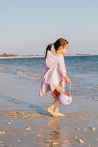 Child in a pink outfit walking on a beach with a sand bucket.