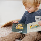 Child holding a book with illustrations of people and text, sitting on a soft surface.